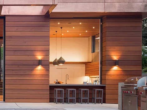 Pass-through window open in a timber-clad wall between an indoor kitchen and an outdoor bar counter with stools