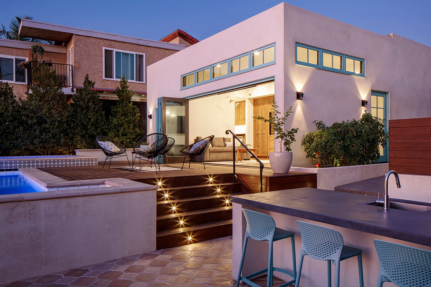 Stucco home at dusk with its glass doors slid open onto a poolside deck and outdoor kitchen